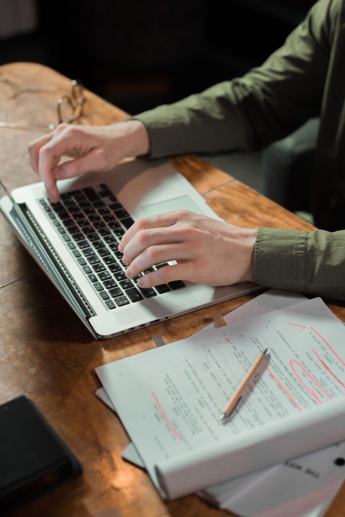 Focused person typing on laptop with notes and papers beside on wooden table.